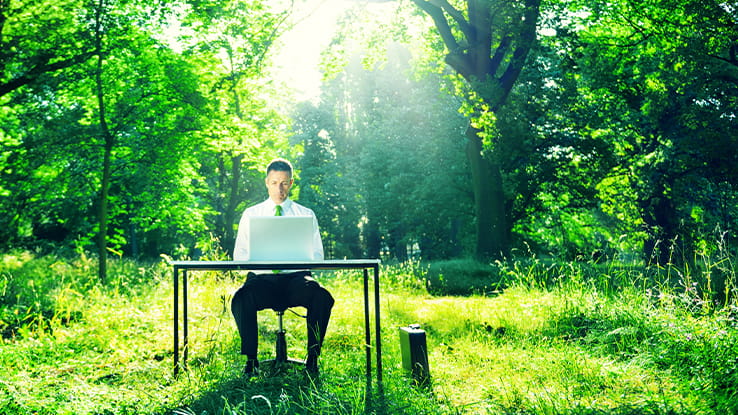 man in pak zit achter bureau met laptop in een groene omgeving met gras en bomen
