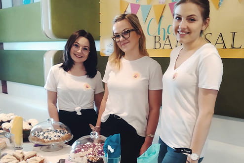 Charity Bake w biurze Colliers. Trzy pracownice Colliers sprzedające ciasta na charytatywny cel.